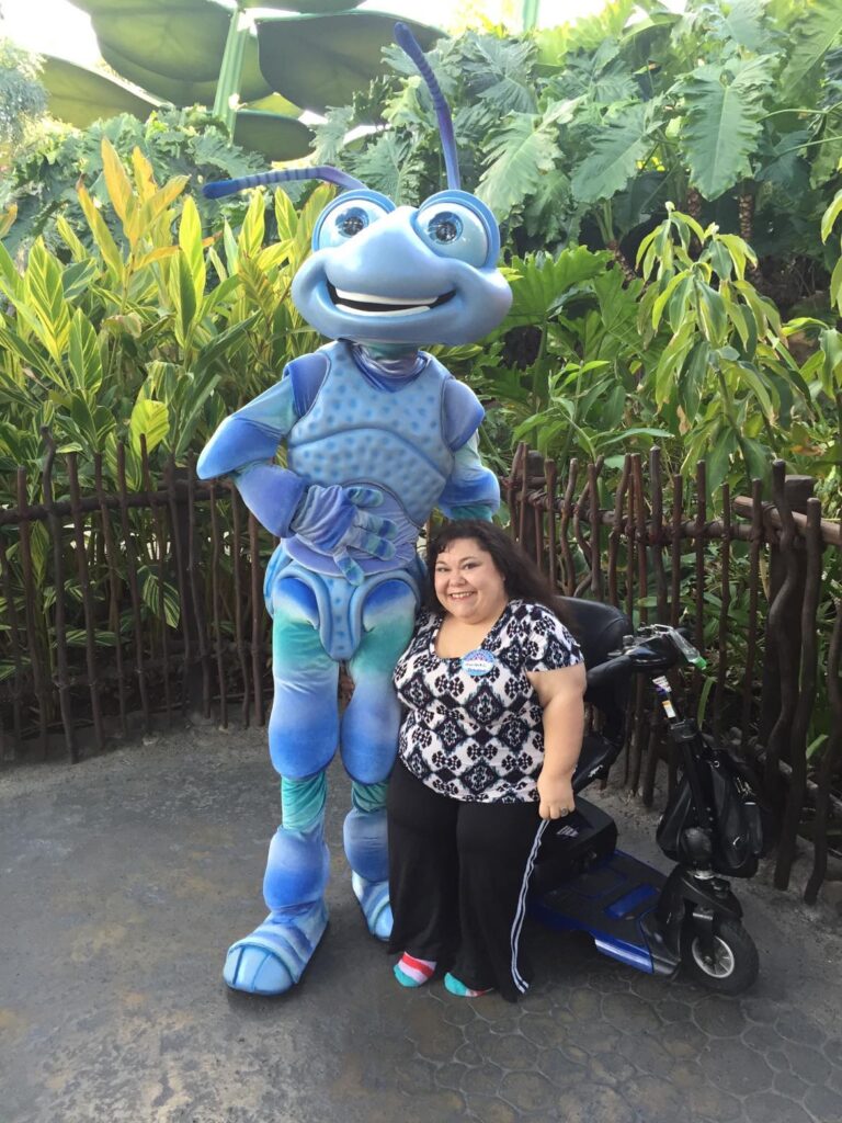 Adult with SEMD. She is next to a huge statue of a bug. She is using a small electric scooter as a mobility device.