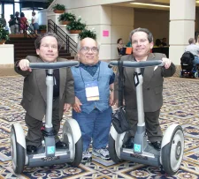 Three men with SEDC. Man in the middle is standing. The men on both sides are identical twins in identical suits riding riding mobility devices.